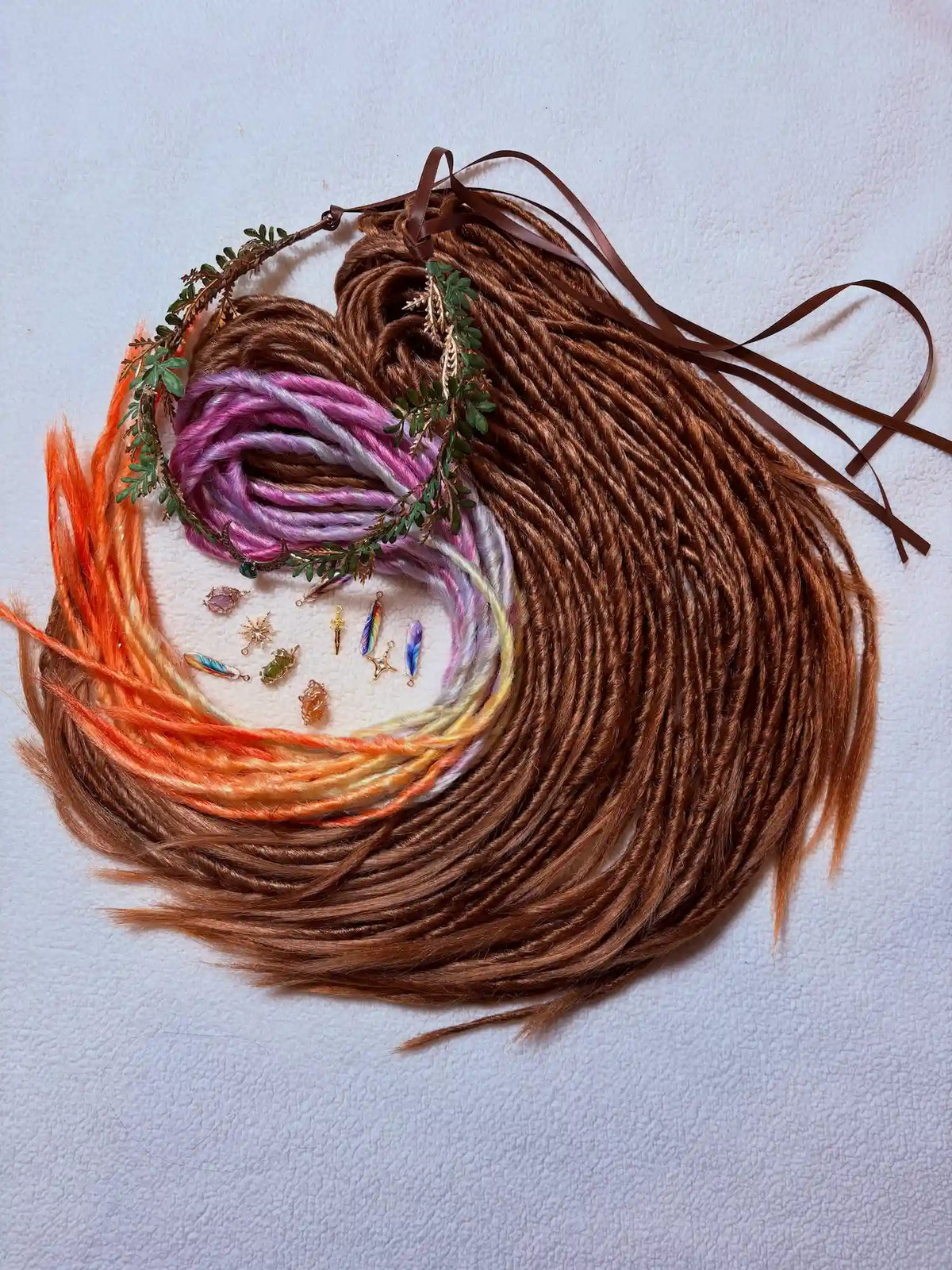 A heart-shaped arrangement of multiple dread extensions in shades of brown, orange, pink, and cream, decorated with small dried flowers and greenery, displayed on a light textured background.