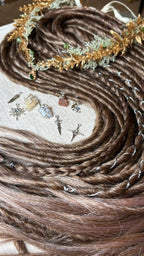 A close-up of a brown dreadlocks with twisted rope-style strands, arranged on a light fabric background with small decorative hair charms and a gold-toned floral hair accessory.