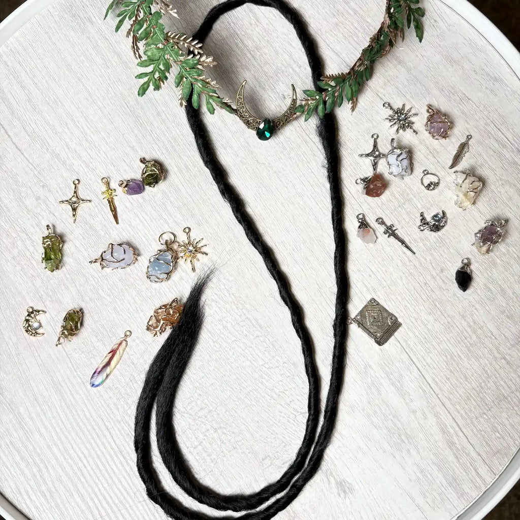 A black dread displayed on a white wood surface, styled with a green leafy crown and surrounded by various small jewelry charms and pendants.